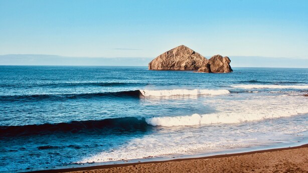 Une plage avec des vagues et une grande formation rocheuse visible au loin.