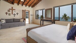 Un dormitorio con cama, sofá, un ventanal con vista al mar y techo de madera.