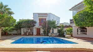 A pool area with a clear blue pool, surrounded by a white building with a tiled roof, palm trees, and a stone wall.