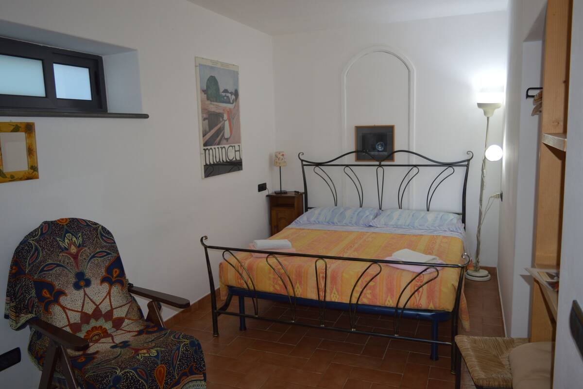 A small, neatly arranged bedroom with a bed, a chair, a bedside table, and a lamp.
