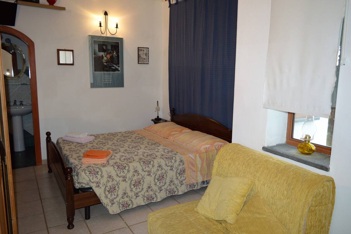 A bedroom with a bed, a chair, a window with a curtain, a wall-mounted lamp, and a small shelf with a decorative item.