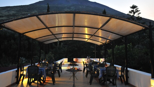 Una terrazza esterna con tavoli e sedie, una zona coperta e una montagna sullo sfondo.