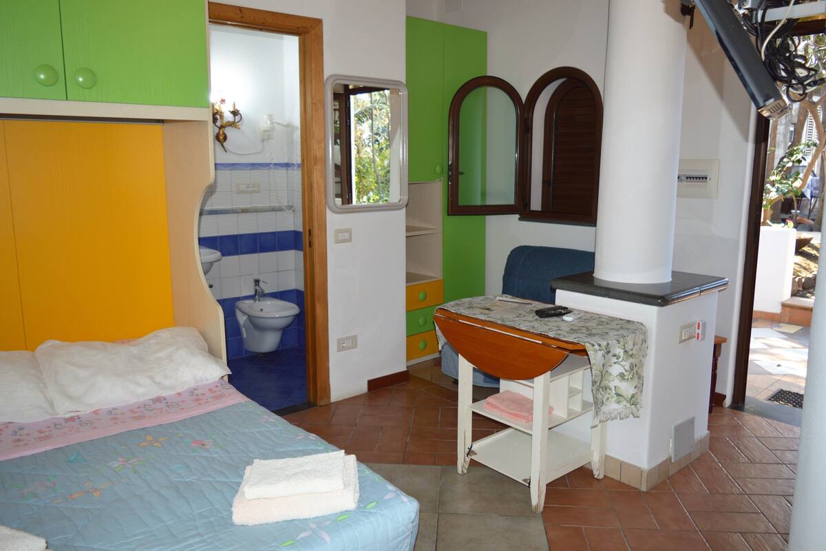 A bedroom with a bed, a small table, a mirror, and a bathroom with a toilet and sink.