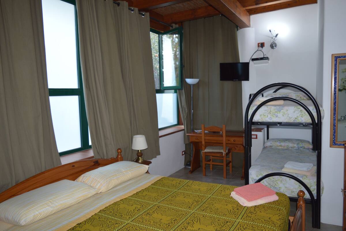 A room with a bed, a desk, and a bunk bed. There is a window with curtains, a lamp, and a flat-screen TV on the wall.