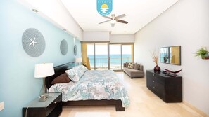 Un dormitorio con cama, mesita de noche, cómoda, ventilador de techo y un amplio ventanal con vistas al mar.