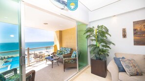 A balcony with a view of the sea, wicker furniture, a potted plant, and a couch with a patterned pillow.