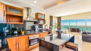 Una cocina moderna con una isla central, horno integrado y microondas. Hay un comedor con vistas al mar.