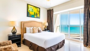 Habitación de hotel con una cama grande, dos mesitas de noche, vista al océano y un cuadro en la pared.