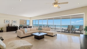 Uma sala de estar ampla com um sofá seccional grande, uma mesa de centro e vista para a praia através de janelas do chão ao teto.