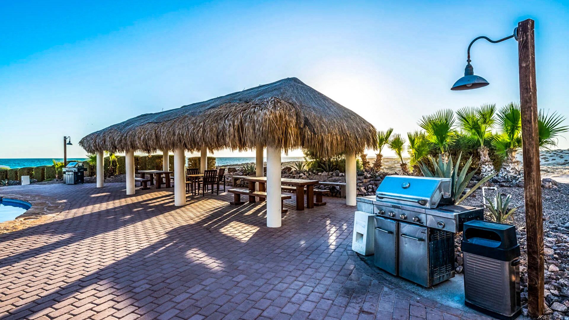 Une terrasse au bord de la plage avec un gazebo à toit de chaume, des sièges d’extérieur, un barbecue et une poubelle.