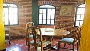 A dining area with a round table surrounded by six wooden chairs with cushioned seats. The room features brick walls and large windows with black frames. - Beautiful house in Chapala with an incredible garden with pool (Chapala)