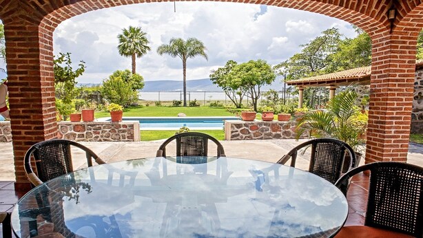 An outdoor patio with a glass table and wicker chairs, overlooking a swimming pool and a mountainous landscape. - Beautiful house in Chapala with an incredible garden with pool (Chapala)