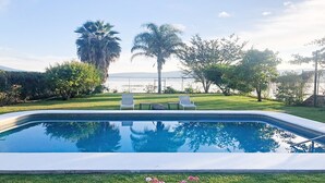 A swimming pool with two lounge chairs, surrounded by well-maintained grass and trees, with a view of the sea and hills in the background. - Beautiful house in Chapala with an incredible garden with pool (Chapala)