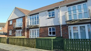 A two-story residential building with a balcony on the upper floor, a brick facade, and a green fence in front. - 2 bedroom accommodation in Beadnell, near Alnwick (Beadnell, near Alnwick)