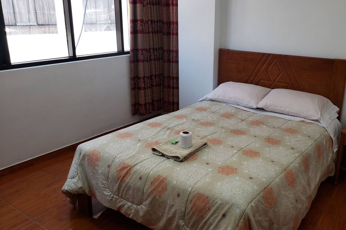 Una cama bien tendida con una colcha estampada, un cabecero de madera y una mesita de noche con un cajón.