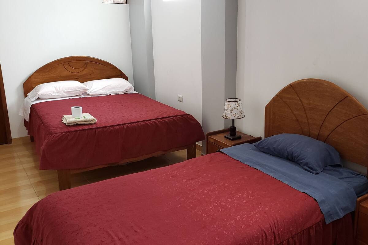 Una habitación con dos camas, cada una con cabecero de madera, colcha roja y almohada azul. Entre las camas hay una mesita de noche con una lámpara.