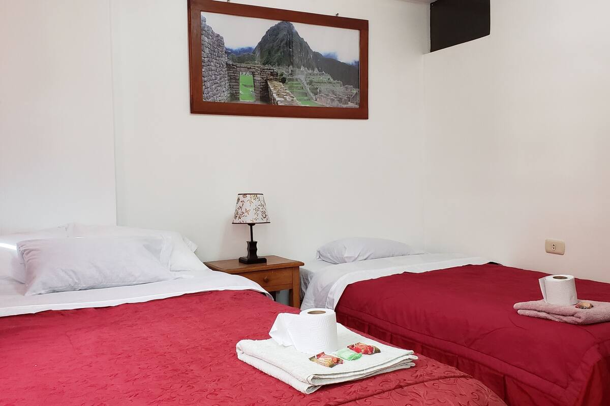 Una habitación de hotel con dos camas, un cuadro de Machu Picchu, una mesita de noche con una lámpara y una cama para gato.