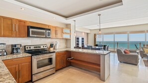 Cocina moderna con encimeras de granito, electrodomésticos de acero inoxidable y armarios de madera. Hay una isla grande con asientos y vistas al océano a través de amplias ventanas.