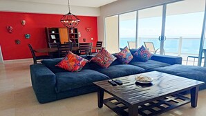 Una sala de estar moderna con un sofá azul, almohadas coloridas, una mesa de centro de madera y un comedor con una pared de acento roja y una araña.