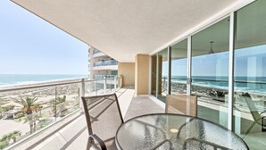 A balcony with a glass table and chairs overlooking a beach and ocean.