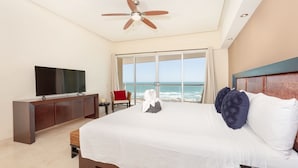 A hotel room with a large bed, a TV on a wooden cabinet, a ceiling fan, and a view of the ocean through large windows.
