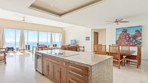 A spacious living area with a large kitchen island, dining table, and chairs. There are large windows offering a view of the ocean.