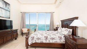 A bedroom with a large bed, a TV, a chair, and a view of the sea.