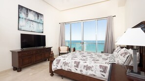 A bedroom with a large bed, a TV on a dresser, a chair, and a view of the sea through a large window.