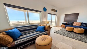 A spacious living room with a large window overlooking the beach, a sofa with patterned cushions, a coffee table, and a side table with a lamp. - Fully Remodeled, The Suite at Rocky Point 2 Ocean front (Puerto Penasco)