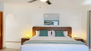 Un dormitorio con cabecera de madera, cama con ropa de cama blanca, dos lámparas de noche, un ventilador de techo y una obra de arte colgada en la pared.