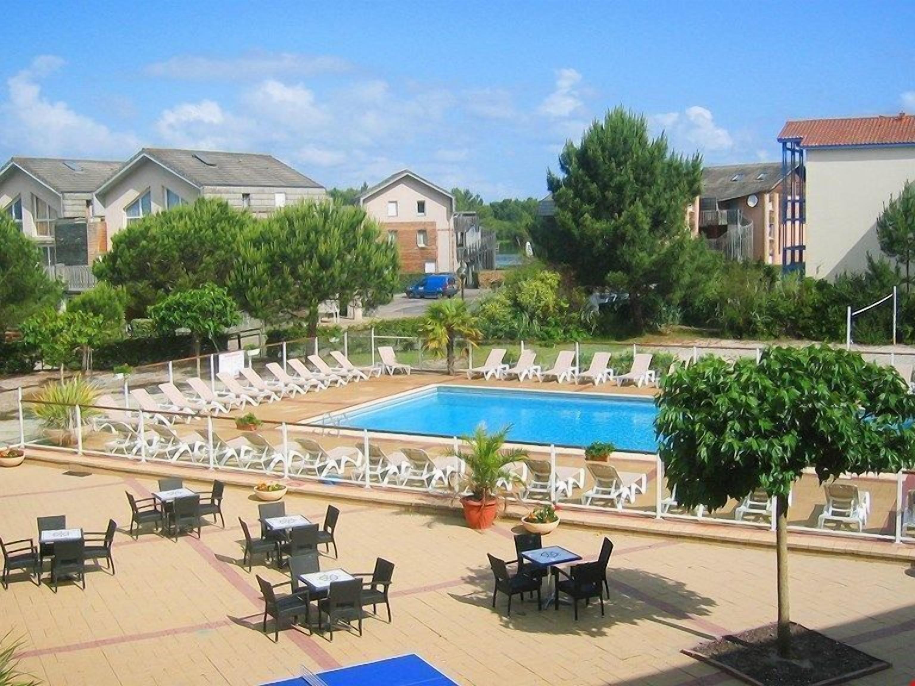Zona de piscina con sillones reclinables y mesas, rodeada de vegetación y edificios.
