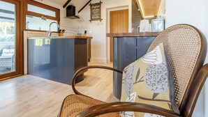 A modern kitchen with a central island, wooden ceiling beams, and a dining area with a wicker chair. - Serenity Sands - UK30265 (Goodrington, near Paignton)