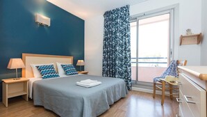 A bedroom with a bed, nightstands, a chair, a dresser, a window with curtains, and a view of the outside. - Appartement Abordable au Bord du Lac (Hourtin)