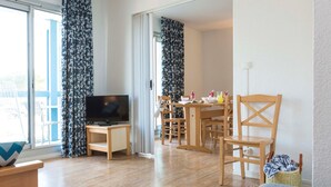 A modern living room with a TV, wooden furniture, and a dining area with chairs and a table. - Appart Duplex à Lac Hourtin | Accès Piscine (Hourtin)