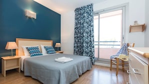Ein Schlafzimmer mit Bett, Nachttischen, einem Sessel, einer Kommode, einem Fenster mit Vorhängen und Blick nach draußen.