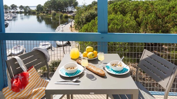 Un balcon avec une table dressée pour le petit-déjeuner, donnant sur une marina où l’on peut admirer des bateaux et la verdure.
