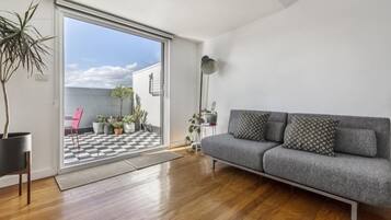 Un salon moderne avec un canapé gris, un plancher en bois et une vue sur un balcon doté d’un sol à carreaux et de plantes en pot.