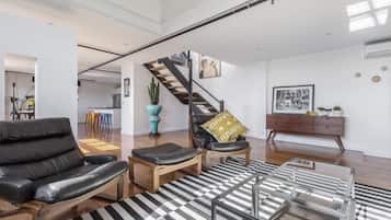 Un salon moderne avec un canapé en cuir noir, une table basse en bois et un tapis à rayures. Un escalier avec une rampe en bois mène à l’étage supérieur.