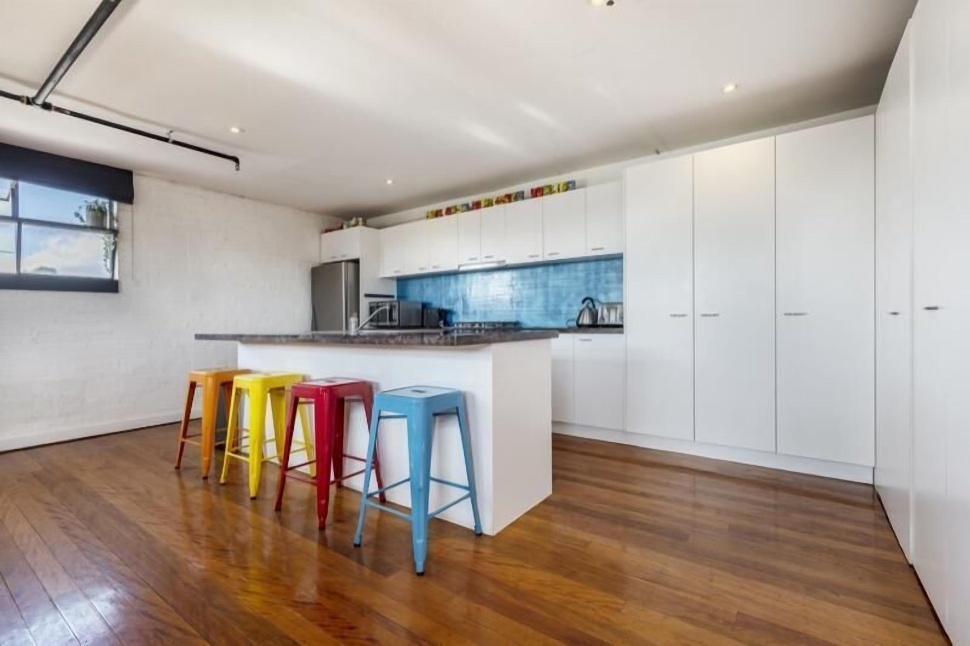 Una cocina moderna con una isla central, armarios blancos y taburetes coloridos.