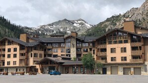A multi-story hotel with a mountain backdrop, featuring balconies, a ski lift, and a parking area.