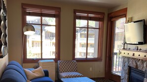 A cozy living room with a blue sofa, patterned armchair, and a wooden dining table. There are large windows with blinds, a fireplace, and a mounted TV.
