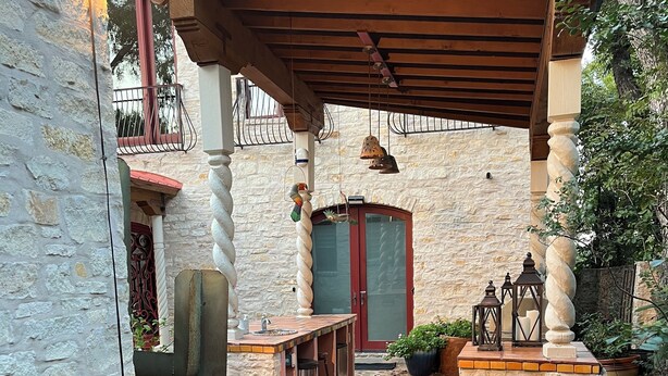 Un sentiero in pietra conduce a un patio coperto con un tavolo e sedie di legno, una porta rossa e piante in vaso.