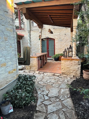 Un chemin de pierre mène à un patio abrité, où se trouvent une table et des chaises en bois, une porte rouge et des plantes en pot.