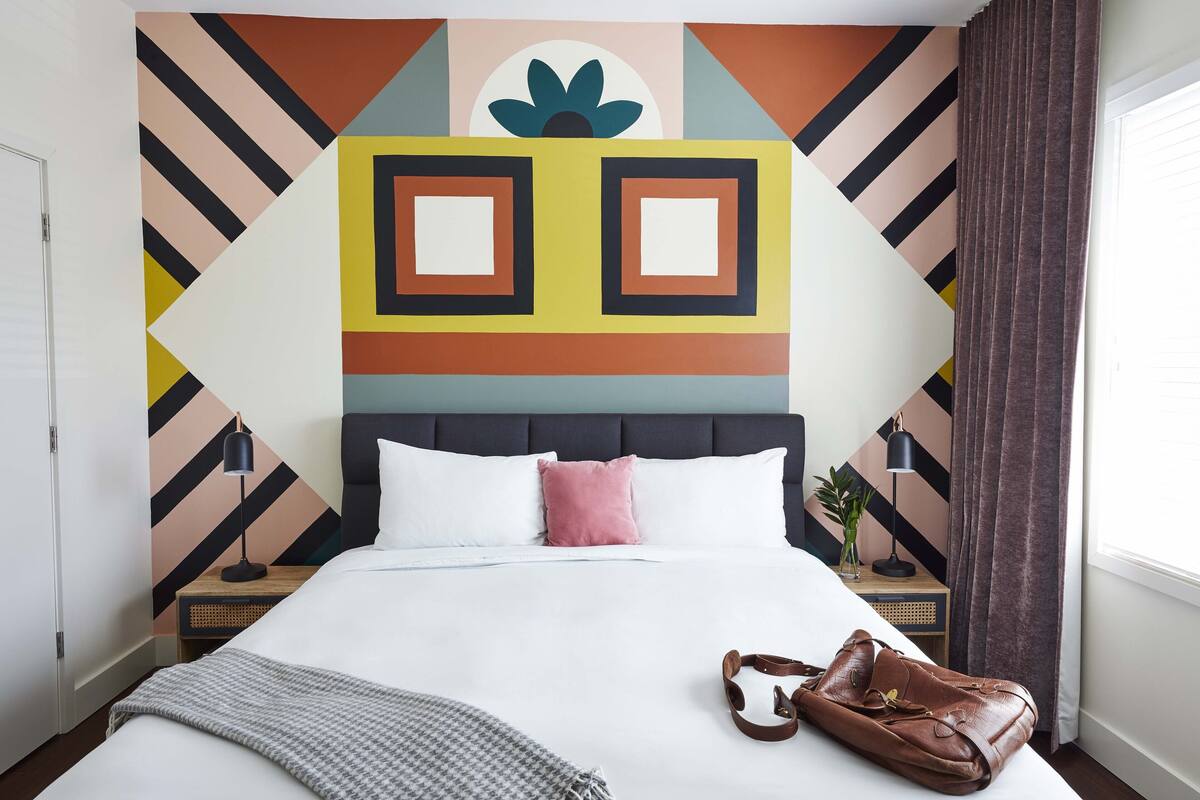 A neatly made bed with a brown leather bag, a headboard with a geometric pattern, and a bedside table with a lamp.