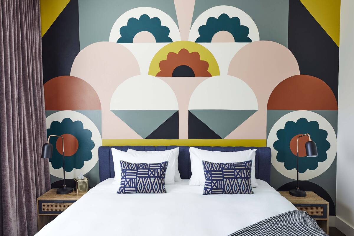 A modern bedroom with a geometric-patterned headboard, a large bed with patterned pillows, and a nightstand with a lamp.