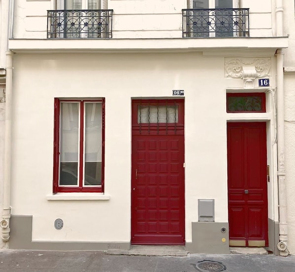 Un bâtiment avec une porte rouge, une fenêtre à cadre blanc et un balcon orné d’une balustrade en fer forgé.