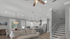 Un salon moderne avec un canapé beige, une table basse en bois et un coin repas comprenant une table et des chaises en bois. On aperçoit à l’arrière-plan une cuisine avec des armoires blanches et des appareils électroménagers en acier inoxydable.