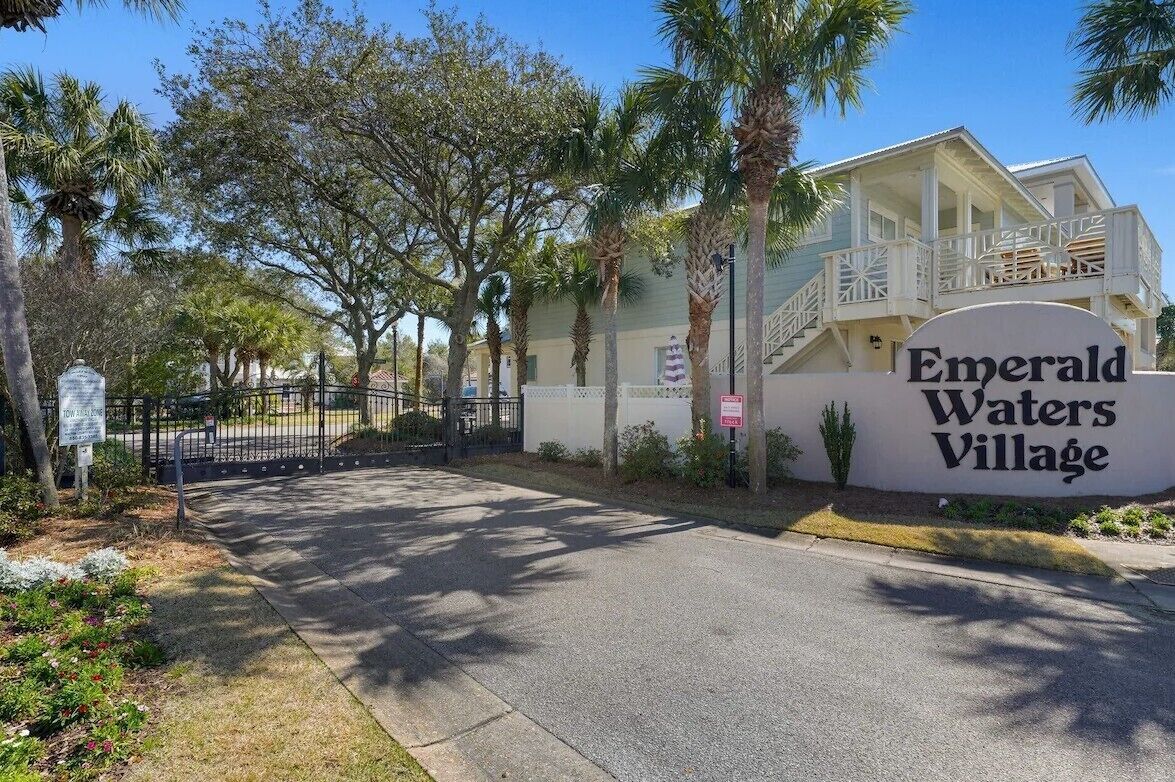 A gated entrance to Emerald Waters Village with a sign, palm trees, and a building with a balcony.