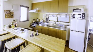 A kitchen with wooden cabinets, a white refrigerator, a microwave, and a sink. There are stools and a counter with various kitchen items. - Nestle Tokyo Deluxe Akihabara 05 (Tokyo)