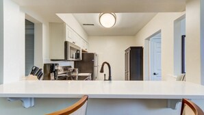 A modern kitchen with white cabinets, a microwave, and a refrigerator. There is a white countertop and a chair in the foreground.
